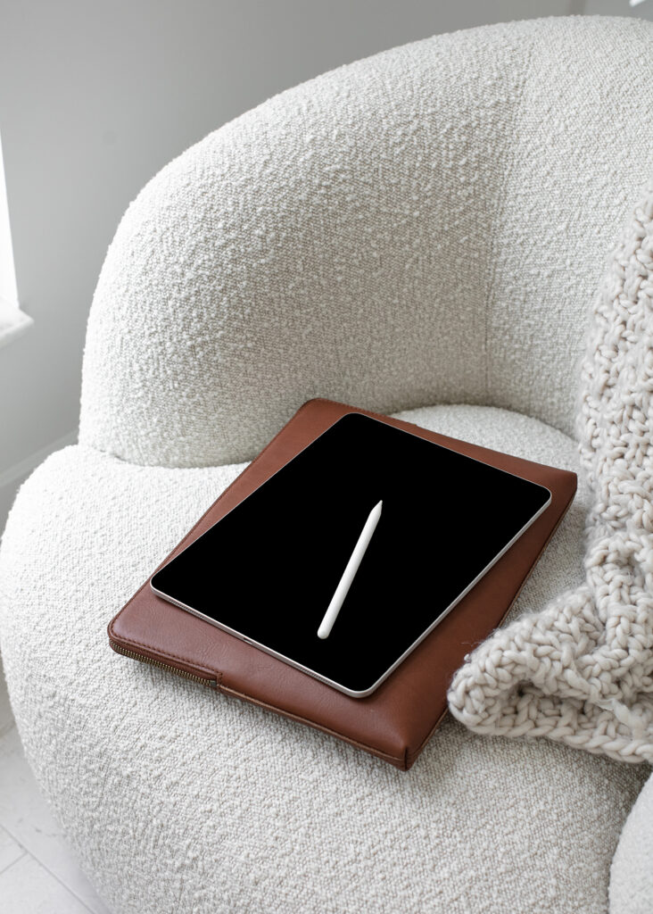  iPad with a white Apple Pencil resting on top of a brown leather book placed on a white armchair.