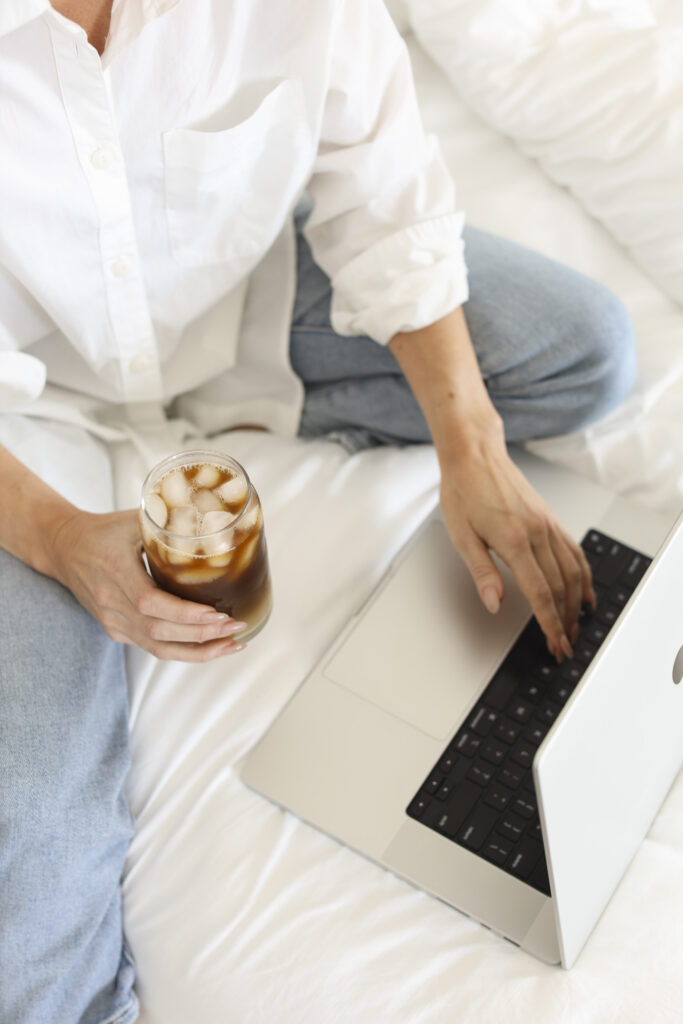 woman wearing white holding a glass of iced coffee and typing on the laptop