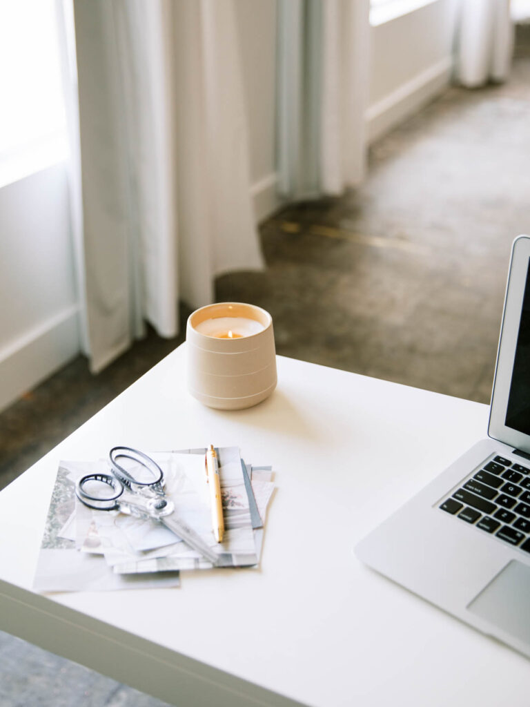 Lit white candle on a wooden table with a laptop, scissors, a pen, and photos scattered around.