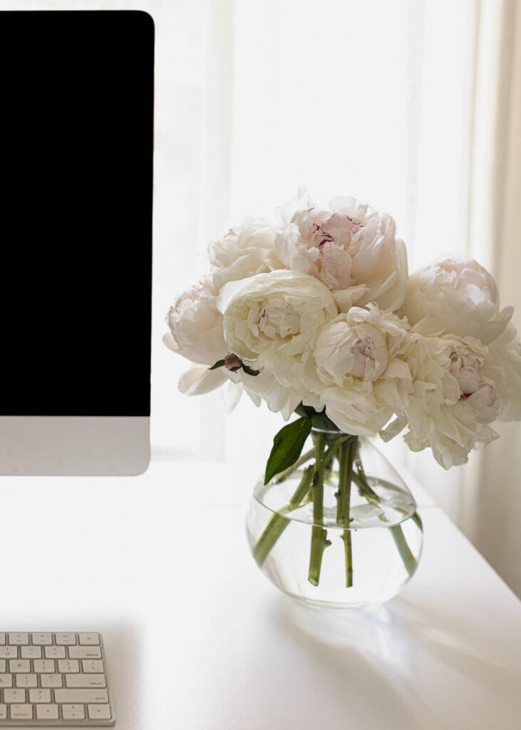 Bouquet of white peonies in a glass vase on a desk next to a computer.